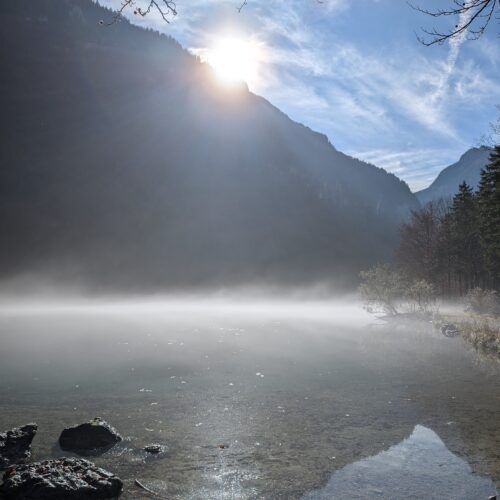 See im Nebel vor einem Berg hinter dem die Sonner hervor sieht.